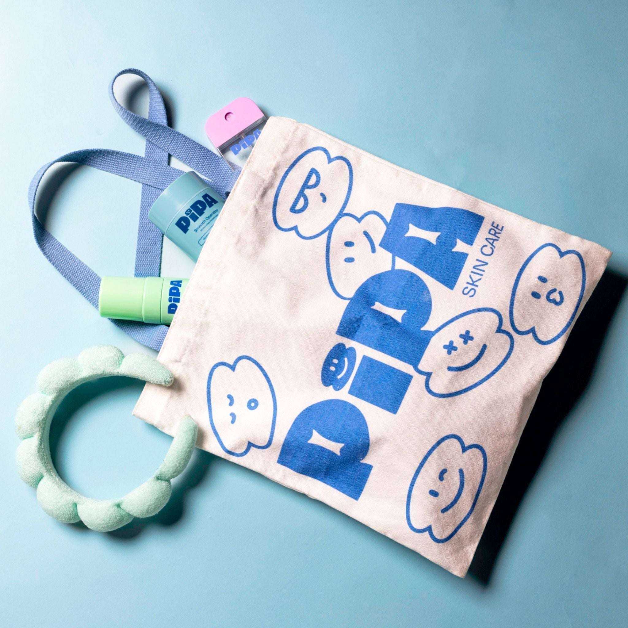 Pipa Tote bag with blue handles and graphic print, surrounded by skincare items.