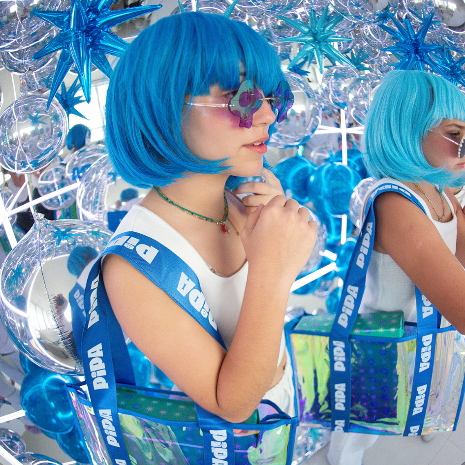 Model with blue wig wearing Pipa Jelly Tote Pipa Blue Bag in a decorative reflective setting