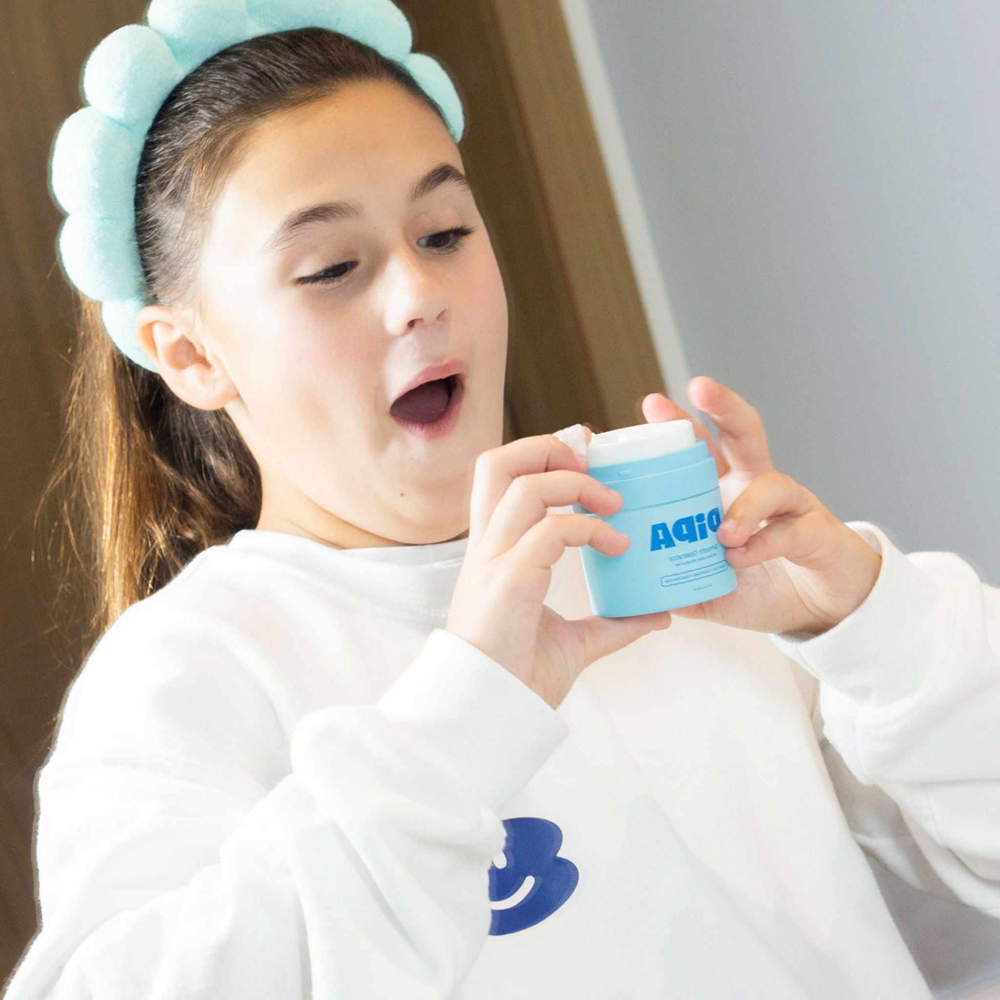 Girl excitedly holding skincare product, wearing blue headband from Daily Glow Bundle for tweens.